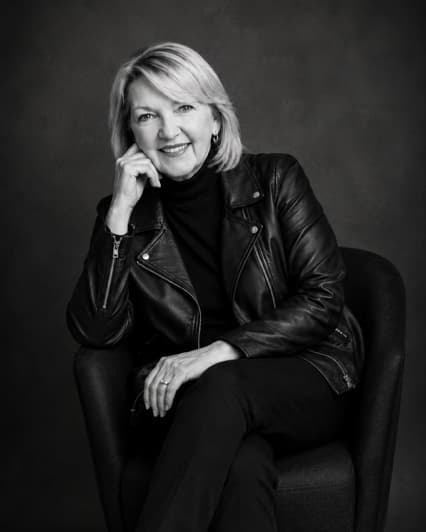 GeorgeAnn Dingus in a black leather jacket, seated with her chin resting on one hand and smiling, in a black-and-white studio portrait