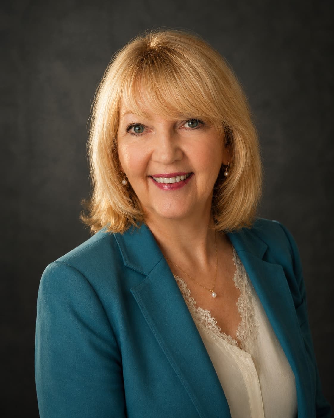 GeorgeAnn Dingus in a teal blazer and pearls, smiling warmly against a dark studio backdrop